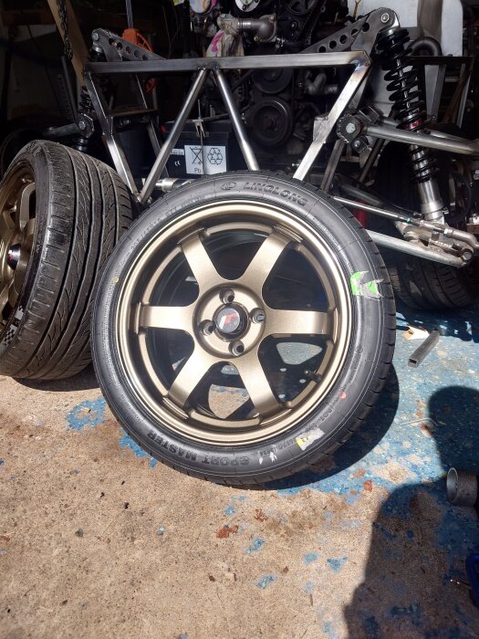 Bronsfärgad bilfälg monterad på ett fordon i en verkstad, med synlig fjädring och motor i bakgrunden.