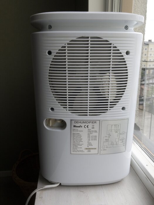 Avfuktare med ventilationsgaller och specifikationsetikett, placerad på ett fönsterbräde. Kabeln syns hängande ner från botten av apparaten.