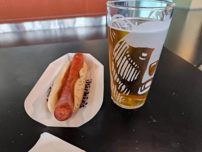 Korv i bröd på ett fat bredvid ett glas öl med björnmotiv, placerat på ett bord vid Helsingfors flygplats.