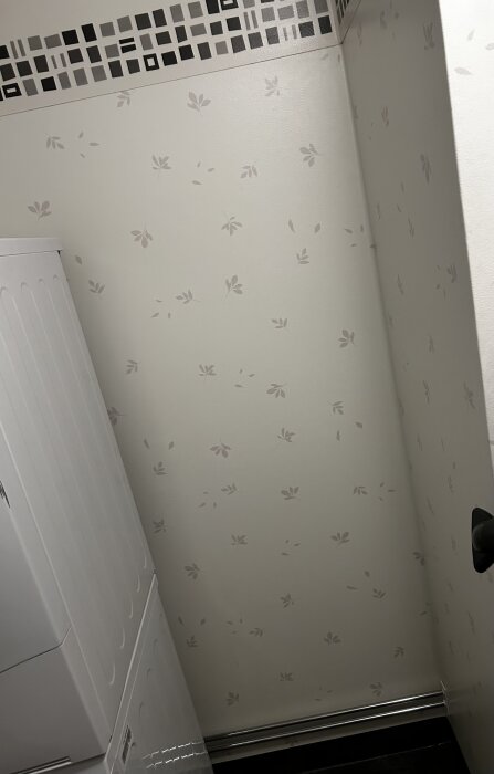 Vägg med ljus våtrumstapet med bladmönster och en svartvit bård högst upp i badrum/tvättstuga, bredvid en vit maskin.