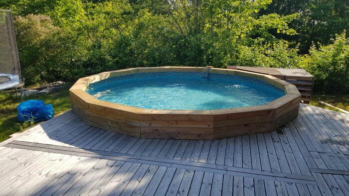 Rund ovanmarkspool med träomgivning på altan, omgiven av grönska. En poolpumphus syns i bakgrunden.