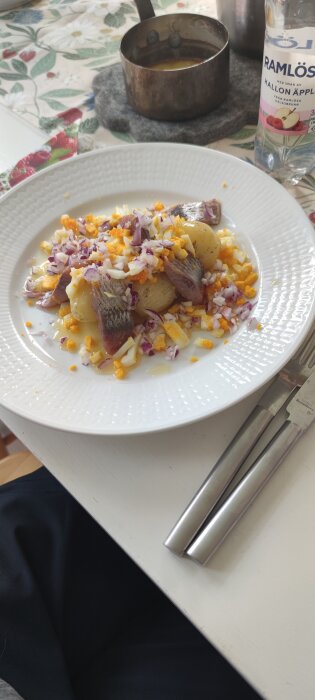 Tallrik med sill, potatis, rödlök och hackat ägg på ett bord bredvid en Ramlösa-flaska och en kastrull med sås.