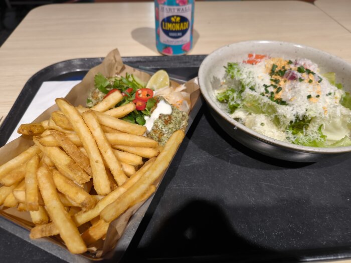 Tallrik med pommes frites, kyckling med koreansk sås och sallad med parmesan på ett matbricka, samt en burk limonad i bakgrunden.