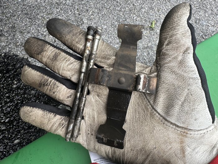 En arbetshandske håller en rostig metallpinne och bromsokshållare över en asfaltbeläggning.