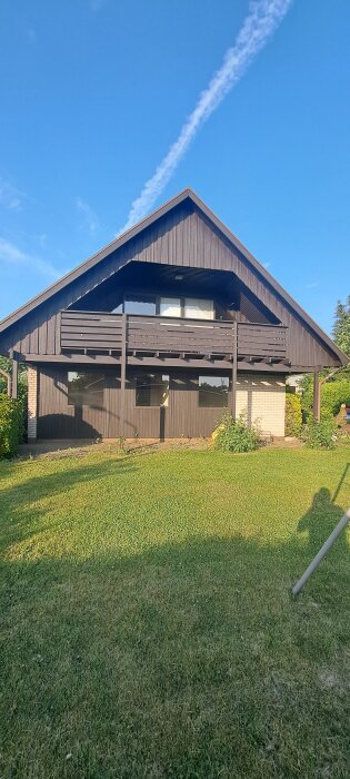 Balkong på en 70-talsvilla med slitna brädor och behov av renovering, sett från trädgården en solig dag med klarblå himmel.