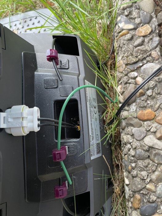 Kablar med klämkontakter kopplade till en robotgräsklippare, omgivna av gräs och sten.