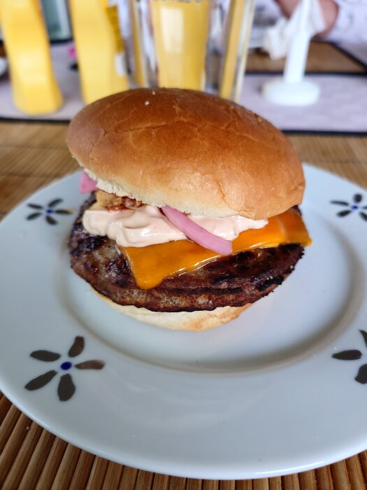 Hamburgare på tallrik med potato bun, Väddö cheddar, picklad och rostad lök, samt sås. Bakgrunden visar ketchup- och senapsflaskor.