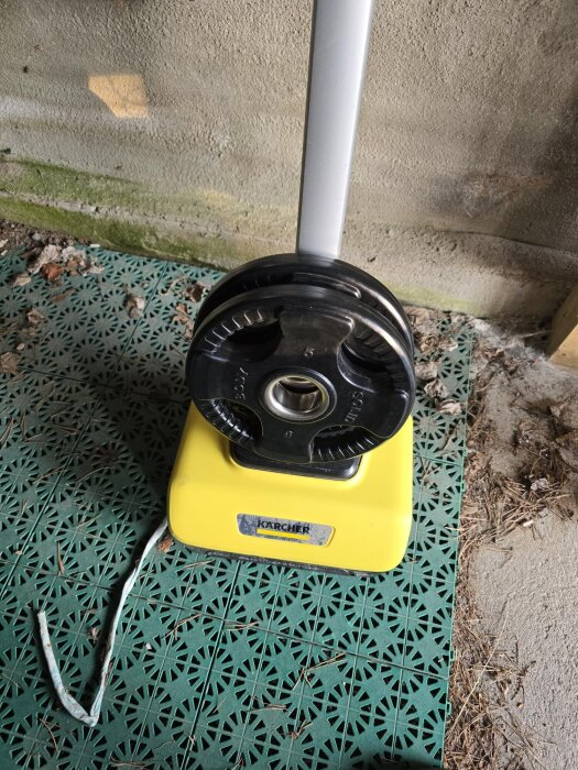 Gul Kärcher-maskin med vikter fästa, stående på grönt underlag bredvid en vägg, med ett vitt spännband liggande på marken.