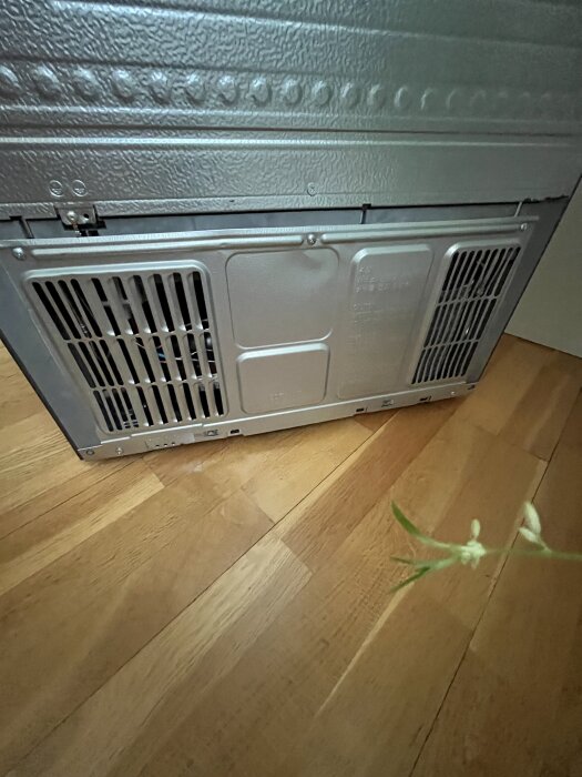 Metalliskt ventilationsgaller vid nedre delen av en hushållsapparat, placerad på ett trägolv med en suddig växt i förgrunden.