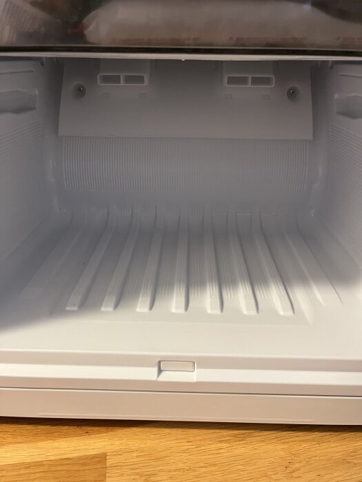 Inuti en tom, öppen frys med ribbad botten och ventilationsöppningar i bakre väggen, sedd ovanifrån på ett trägolv.