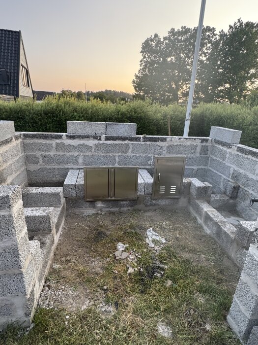 Stenmur under uppbyggnad med två inbyggda metallskåp i en trädgårdsmiljö vid solnedgång.