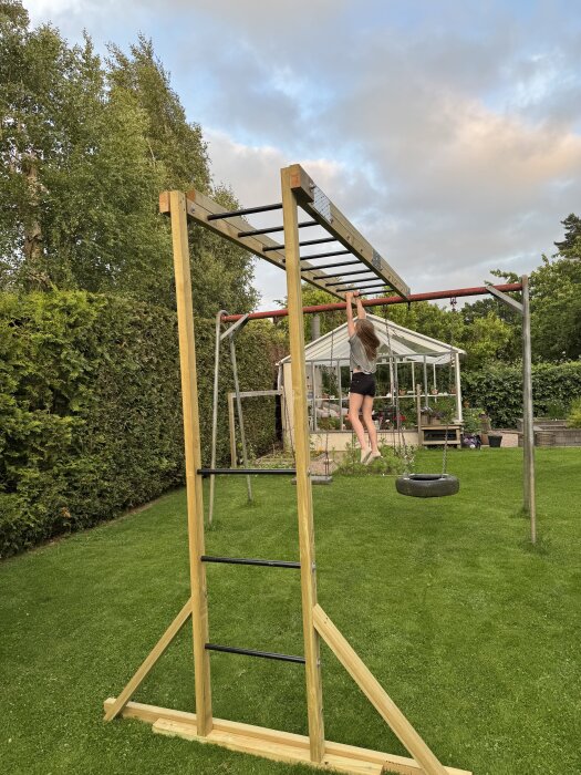 Träställning i trädgård med förstärkta reglar och hålplattor. Person hänger i ställningen. Grönska och växthus i bakgrunden.