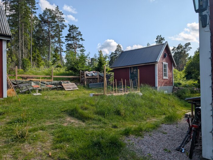 Rött litet hus ovanpå jordkällare med gräsmatta och staket i bakgrunden, omgiven av skog och träd, klar himmel.
