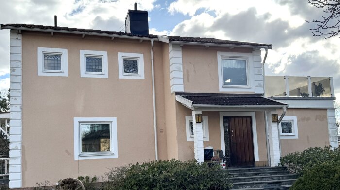 Tegelfärgat tvåvåningshus med vita detaljer och brutet tak. Entré med träport och trappsteg. Kakelplattor och glasräcke syns på övervåningen.