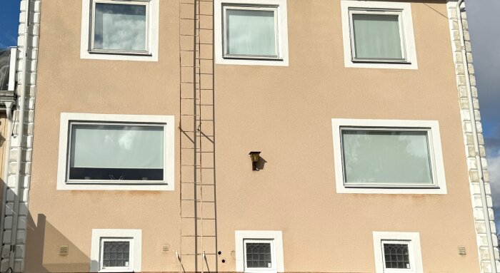 Fasad av ett beige hus med fyra fönster, en stege och en vägglampa.