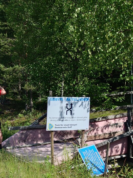 Skylt i skogsmiljö som visar instruktioner för skidåkare att byta sida. En annan skylt lutar mot en trästruktur i bakgrunden.