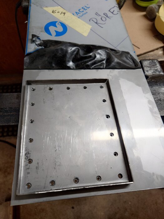 Rostfri stålplåt med förborrade hål, omgiven av skyddsplast och märkningar.