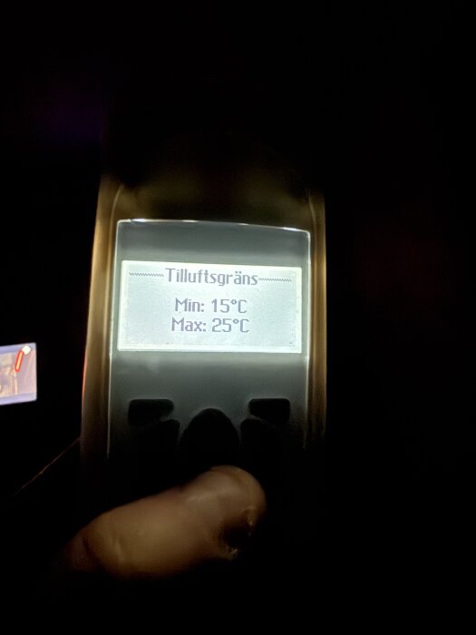 Hand som håller en termometer med skärmen som visar "Tilluftsgräns", Min: 15°C, Max: 25°C, i en mörk omgivning.
