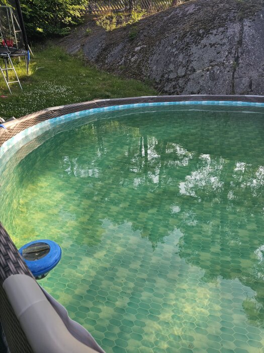Grönskimrande pool med grönaktigt, genomskinligt vatten omgivet av grönska och berg, med ett blått pooltillbehör synligt vid kanten.