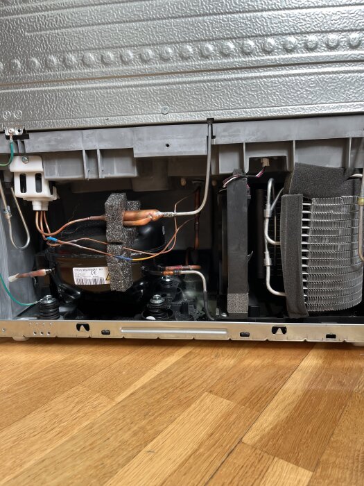 Inre delen av en apparat visar elektriska kablar, kompressor, fläkthjul och metallrör på en trägolvsyta. Inga tecken på avdunstning eller gummipropp.