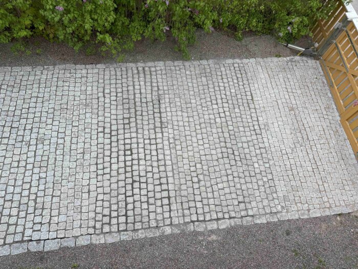 Bild av stenläggning framför garage; ojämt mönster och olika sandfärger syns, med kantstenar i varierande höjd mot gatstenen.