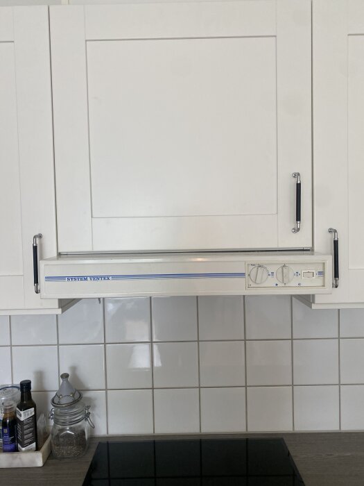 Inbyggd köksfläkt under vita köksskåp, omgiven av kakelvägg och köksbänk med flaskor och burk.