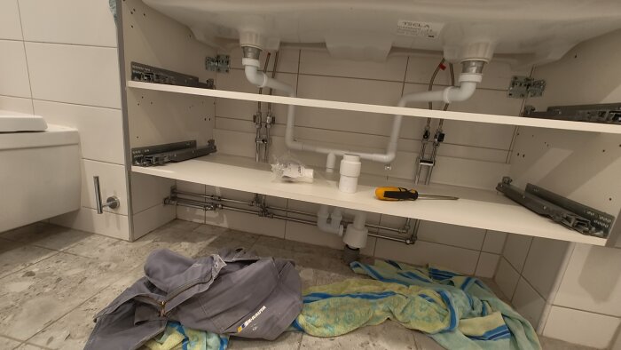 Två handfatsinstallationer med rör och avlopp under, verktyg och en handduk på golvet, samt en vakuumventil synlig på hyllan.