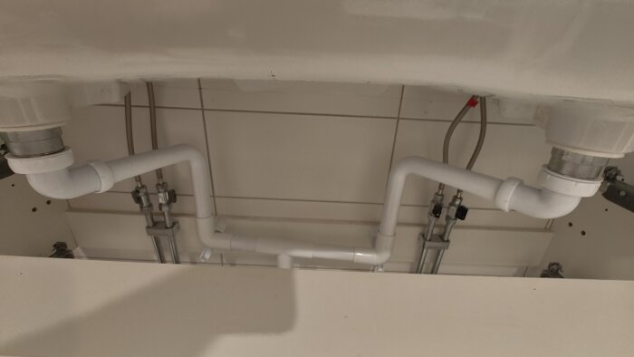 Avloppsrör under handfat där flera 90-gradersböjar kopplar samman ledningar mot väggen, med vita PVC-rör och beslag.