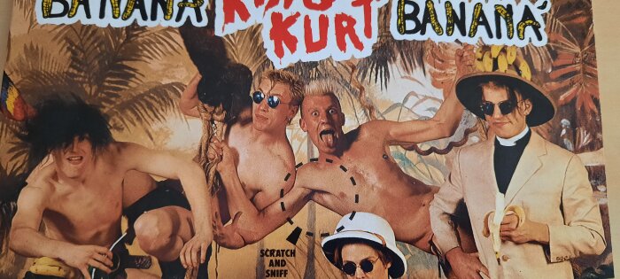 Fem personer poserar framför en tropisk bakgrund. En man i kostym håller i en banan. Texten "scratch and sniff" syns.