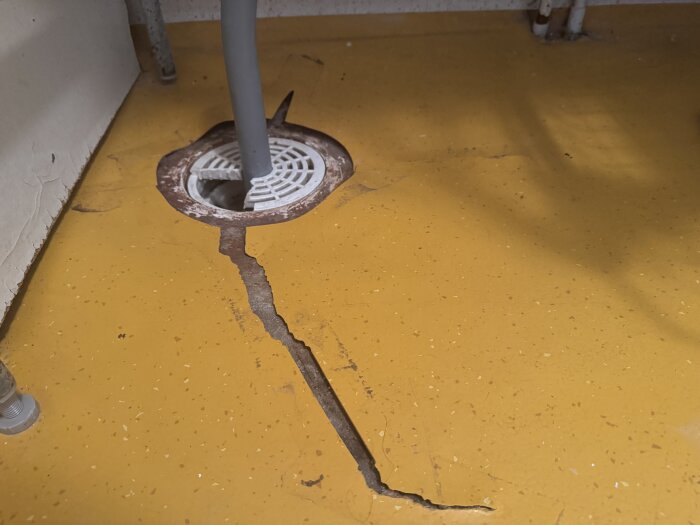 Gul plastmatta i källarens tvättstuga med sprickor runt avloppsbrunn; upphöjda kanter syns.