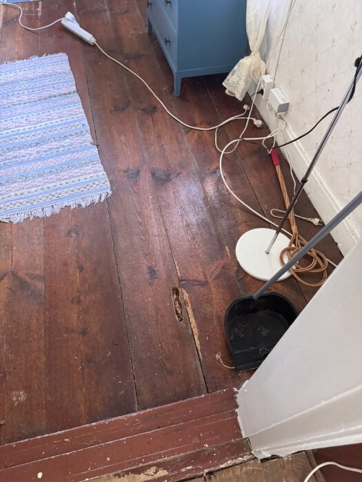 Trägolv med ett avlångt hål, möjligen orsakat av skadedjur som husbock. En blå byrå och sladdar syns i bakgrunden.