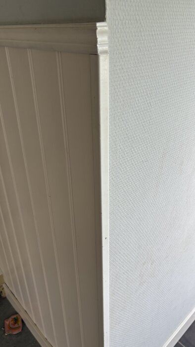 Ett hörn med ljus panel på ena sidan och vit strukturtapet på den andra.