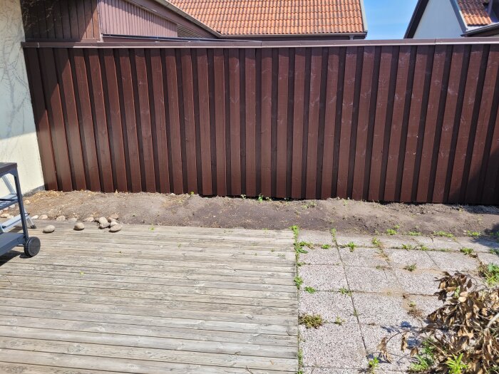 Trädgård med trädäck, marksten, brunmålat trästaket, och plan markyta för framtida odlingslådor, grill och pizzaugn.