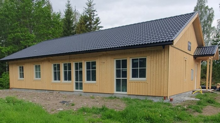 En träbyggnad med svart tak och många fönster på en nästan ofärdig tomt, omgiven av grönska och träd.