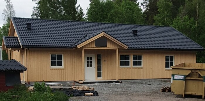 En nybyggd träfasad hus med svarta takpannor och två fönster, omgiven av grönska och en container på tomten.