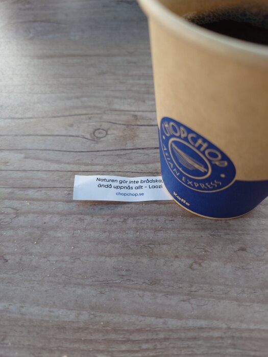 Pappersmugg med kaffe på träbord, citat från Laozi syns delvis på etikett.