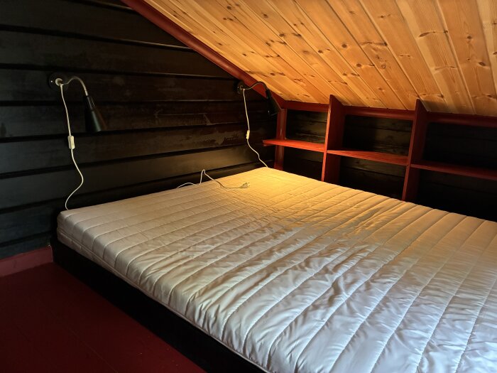Madrass och sänglampor på plats i ett rum med lutande träpaneltak och svarta väggar.