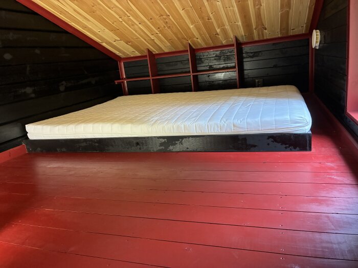 Madrass placerad på röd träplattform med snedtak och mörka träväggar.