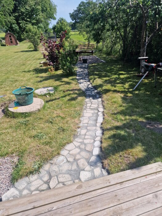 En smal stengång leder genom en grönskande trädgård med buskar och träd, mot en avlägsen uteplats med bänkar.