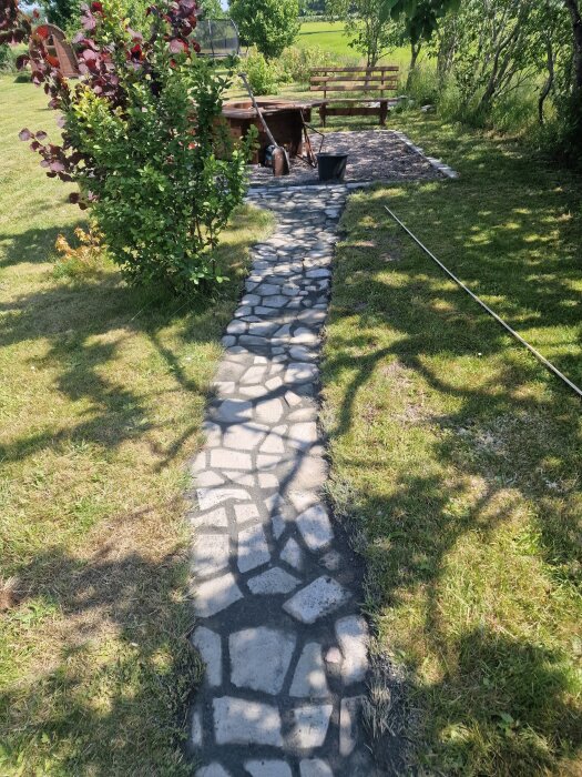 Stengång genom trädgård, omgiven av grönska, leder till en grillplats med bänk. Verktyg och krukor syns vid sidan av stigen.