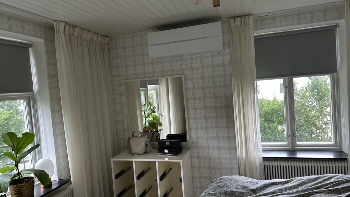 Väggmonterad luftvärmepump ovanför byrå i sovrum med två fönster och vita gardiner.