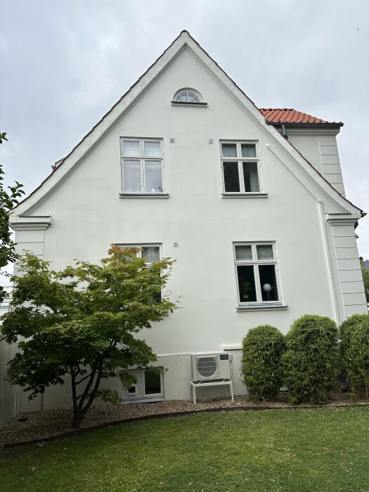 Fasaden på ett vitt hus med brunt tak, fönster och grönska. En luftvärmepump installerad vid marknivå.