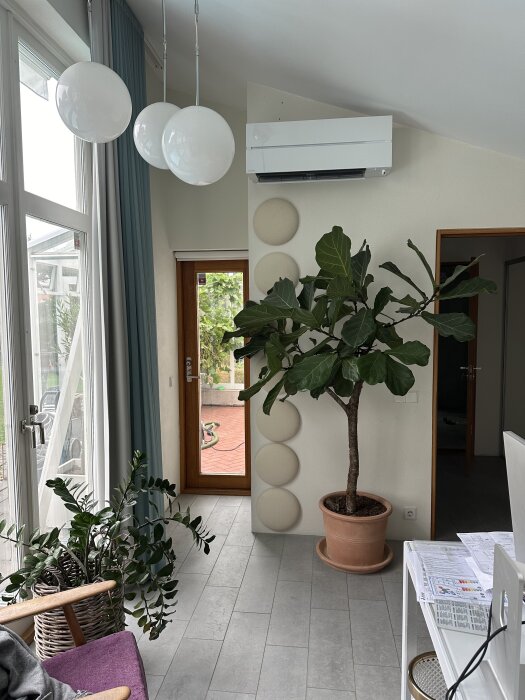 Värmepump installerad på vägg i kontorsrum med stora fönster, gröna växter och hängande lampor.