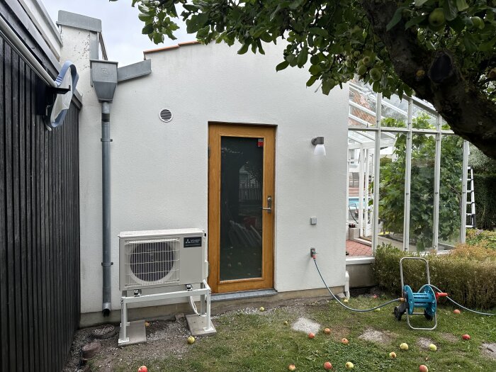 Utomhusdel av en japansk Mitsubishi luft-luftvärmepump installerad på en vit fasad vid sidan av en dörr, med en trädgårdsslang bredvid.