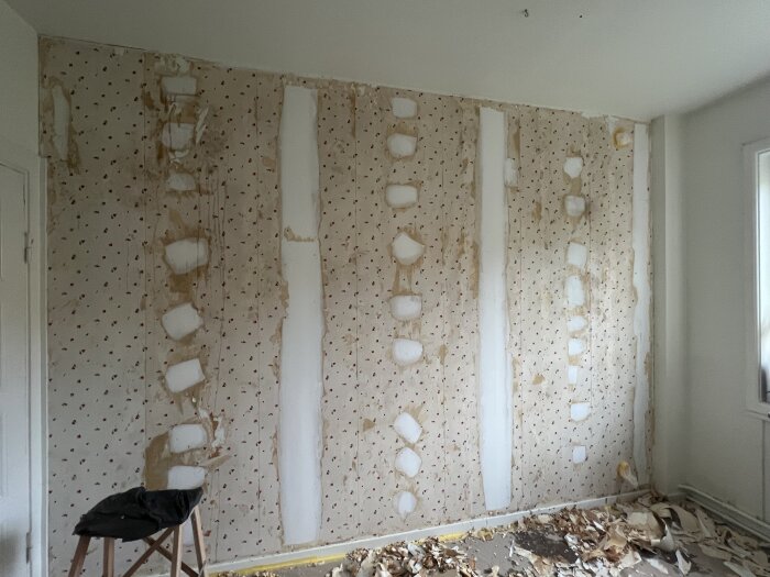 Vägg med borttagen tapet, kvarvarande tapetrester och spackel i fläckar, golvet täckt med tapetrester.