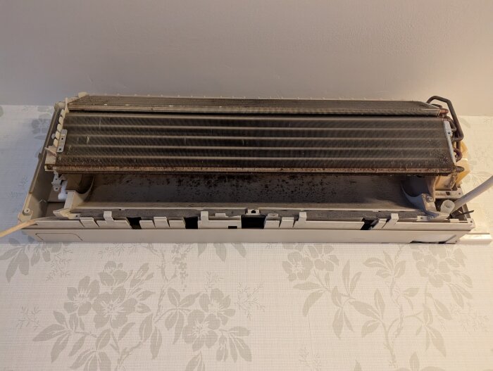 Öppen och smutsig luftkonditioneringsenhet på en blommönstrad yta, i behov av rengöring.