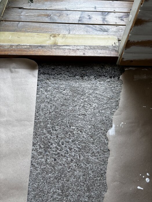 Grovt betonggolv i förråd med papper intill, delvis täckt av trätröskel, där ytan är dammig och ojämn.