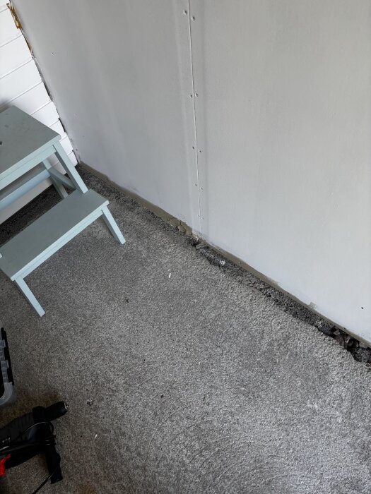 Kraftigt texturerat lagergolv med damm och smuts, grått med en ljus vägg i bakgrunden och en liten blå trappstol bredvid.