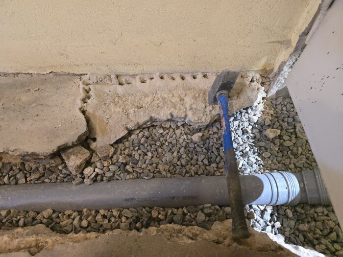 Bruten betongvägg med ett VVS-avloppsrör i grusfyllning och en hammare.