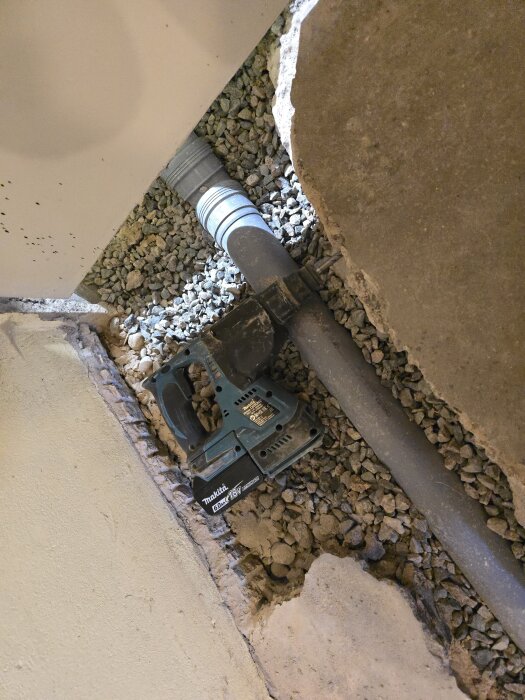 Öppen ränna i hobbyrum, exponerad avloppsledning med grus runt, borr och Makita-verktyg placerade längs sidan, under renoveringsarbete.